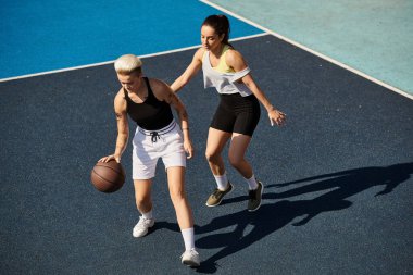 İki genç kadın, arkadaşlar, bir basketbol sahasının tepesinde gururla dururlar, yaz güneşinde güçlü ve sportmence davranırlar..