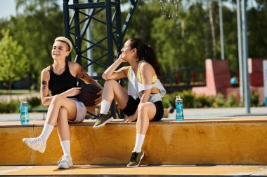 İki atletik genç kadın bir çıkıntıda oturuyor, dışarıda bir yaz gününün tadını çıkarırken bir anlık neşe ve kahkaha paylaşıyorlar..