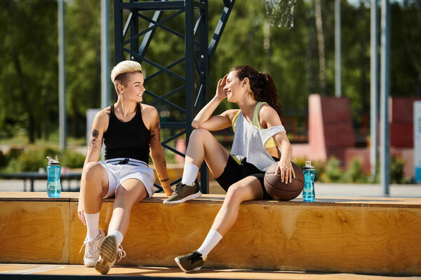 Две молодые спортсменки наслаждаются отдыхом на деревянной скамейке после игры в баскетбол на открытом воздухе летом.