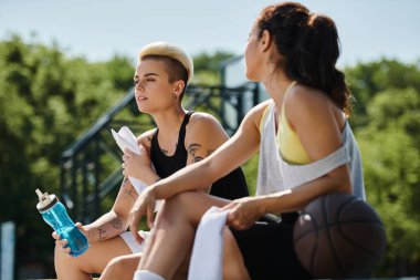 Basketbol sahasında birlikte oturan iki genç kadın, oyuna ara veriyorlar, dostluk ve dostluk gösteriyorlar..