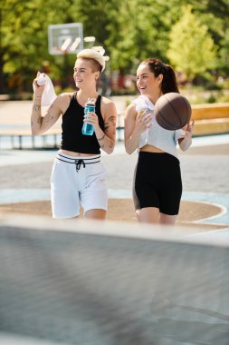 İki atletik genç kadın yan yana duruyorlar, basketbol toplarını açık havada tutuyorlar, güven ve dostluk yayıyorlar..