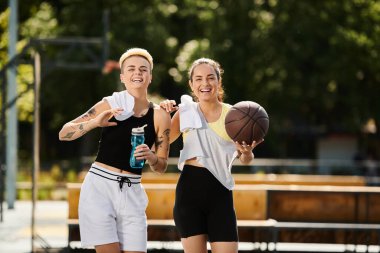 İki genç atletik kadın bir basketbol topuyla yan yana durup güneşli bir havada maçın keyfini çıkarıyorlar..