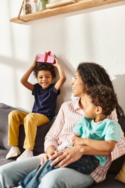 cheerful little african american boy holding present and looking at his mom and brother, Mothers day clipart