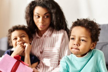 focus on adorable african american kid holding present with his blurred mom and brother on backdrop clipart