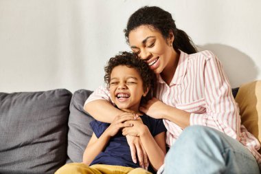 cheerful loving african american mother hugging her adorable smiling son while in living room clipart