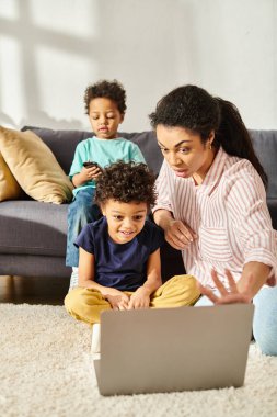 Sevimli Afrikalı Amerikalı çocuk kardeşinin yanında oturuyor ve laptoplu düşünceli anne