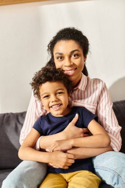 cheerful african american mother and son in homewear hugging and looking at camera, modern parenting clipart