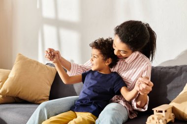 cheerful african american mother holding her little cute son and sitting on sofa in living room clipart