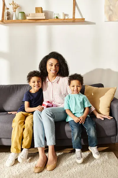 stock image joyous african american mother holding present and sitting on sofa with her sons, looking at camera