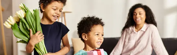 stock image focus on happy african american boys with tulips and gift near blurred mom, Mothers day, banner