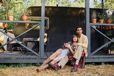 An interracial man and woman sit together on the ground, taking a peaceful break during their romantic getaway in a natural setting. clipart