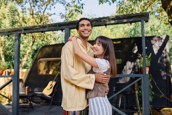 Межрасовая пара тепло обнимается перед палаткой на живописном фоне природы.