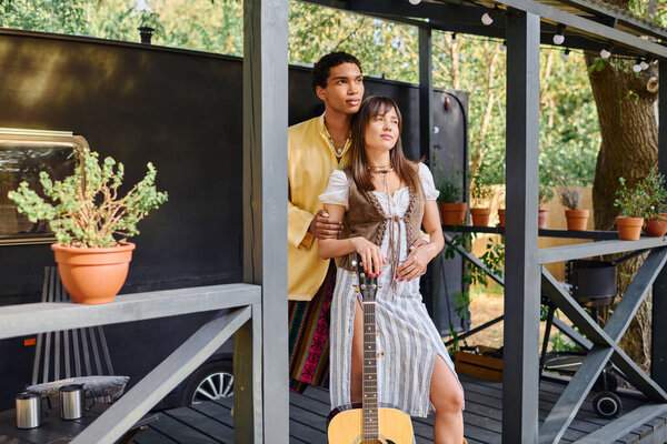 A man and a woman, an interracial couple, stand next to a guitar in a natural setting, enjoying a romantic moment together.
