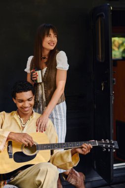 Bir adam bir kadının yanında gitar tutuyor, sakin dağ ortamında birlikte müzik yaratıyor..