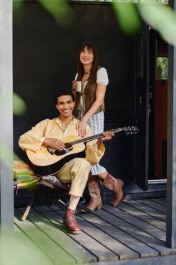 A man and woman sit on a porch playing guitar together, enjoying a moment of musical connection in a serene outdoor setting. clipart