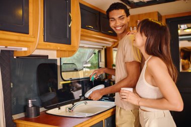 A man and woman, of different ethnicities, stand in a kitchen next to a sink. clipart
