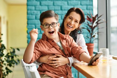 cheerful inclusive preteen boy with Down syndrome smiling at camera while holding phone near his mom clipart