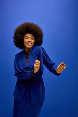 Kıvırcık saçlı, canlı bir arka planda poz veren mavi takım elbiseli şık Afro-Amerikan kadın..