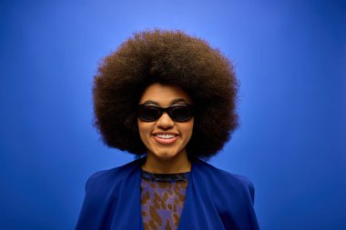 Şık Afro-Amerikalı kadın, parlak arka plana karşı modaya uygun mavi ceket ve güneş gözlüğü takıyor..