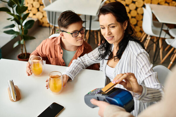 beautiful jolly mother paying with credit card next to her inclusive son with Down syndrome in cafe