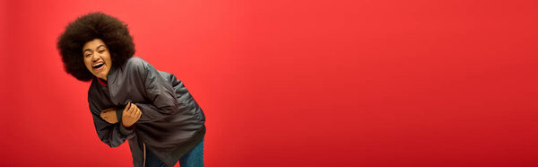 Stylish African American woman with curly hairdohair poses confidently in front of vibrant red wall.