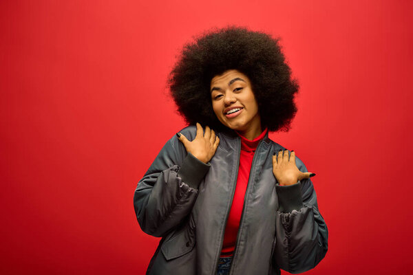 Stylish African American woman with curly hairdo posing in trendy attire against a vibrant red backdrop.