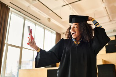 A joyful African American woman in a graduation cap and gown proudly holds up her diploma amidst a celebratory atmosphere. clipart