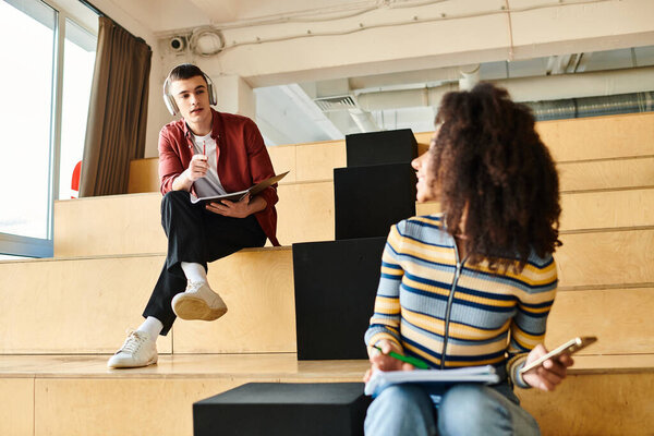A man and woman, both students, engage in a conversation while seated on steps indoors at a university