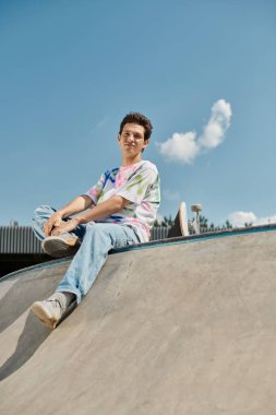 A young skater boy sits calmly at the top of a skateboard ramp in a vibrant outdoor skate park on a sunny summer day. clipart