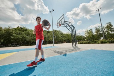 Genç bir adam, güneşli bir günde canlı bir kortta dikilirken kendine güvenerek basketbol topunu tutuyor..