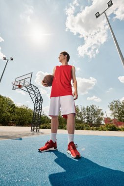 Genç bir basketbolcu elinde basketbol topuyla sahada duruyor ve yaz sıcağında oynamaya hazır..