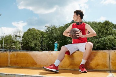 Bir çıkıntıda oturan genç bir basketbolcu, bir basketbol topunu ustalıkla tutuyor..