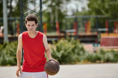 Canlı kırmızı tişörtlü genç bir adam güneşli bir yaz gününde dışarıda basketbol yeteneklerini sergiliyor..