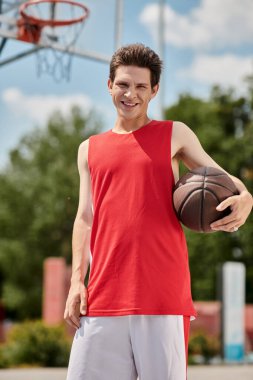 Kırmızı gömlekli genç bir adam güneşli bir yaz gününde basket topunu ustalıkla tutuyor..