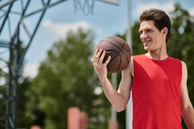 Ateşli kırmızı gömlekli genç bir adam, sıcak yaz gökyüzünün altında atış yapmaya hazırlanan bir basketbolu kavrıyor..
