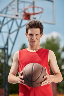 Kırmızı tişörtlü genç bir adam güneşli bir yaz gününde basket topunu ustalıkla dışarı sürüyor..