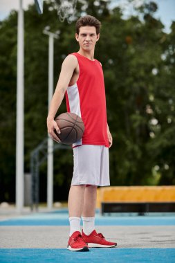 Genç bir adam, güneşli bir günde, açık bir sahada basket oynamaya hazır bir basketbol topunu kendinden emin bir şekilde tutuyor..