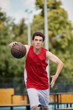 Genç bir adam, canlı bir basketbol sahasının tepesinde, yaz gününün parlak güneşinin altında kendine güvenerek bir basketbol topu tutuyor..