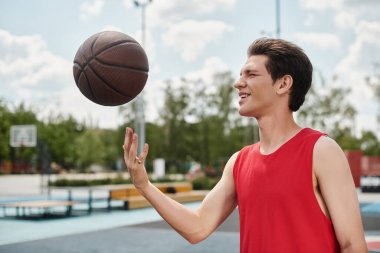 Kırmızı tişörtlü genç bir basketbolcu güneşli bir günde dışarıda basketbol oynuyor..