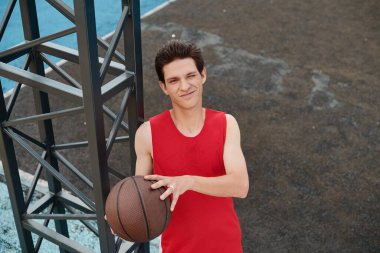Kırmızı tişörtlü genç bir basketbolcunun güneşli bir günde açık havada bir basketbol topunu ustalıkla idare edişini gösteren canlı bir görüntü..