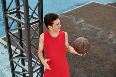 Kırmızı giyinmiş genç bir adam basketbol yeteneklerini bir yaz günü açık havada sergiliyor..