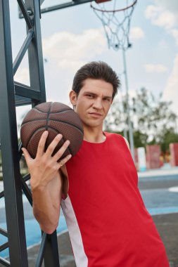 Kırmızı gömlekli genç bir adam bir basketbol topunu ustalıkla tutuyor, bir yaz günü açık havada oynamaya hazır..