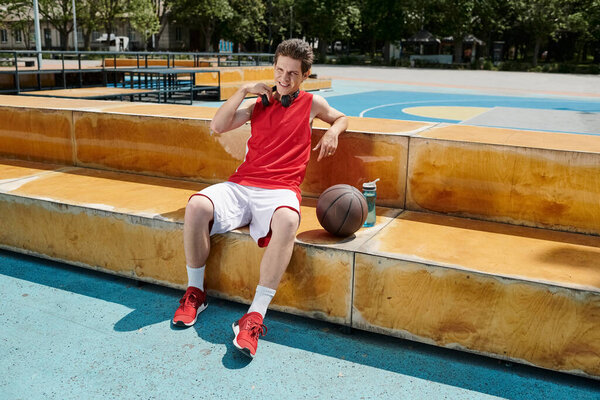A man in a red shirt and white shorts sits on steps with a basketball, showcasing his passion for the game in an outdoor setting.
