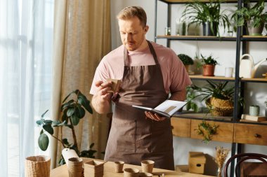 A man in an apron holding pot for plant and notebook in a flower shop clipart