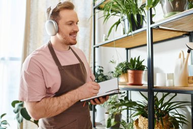 A man wearing headphones stands in front of a shelf in a plant shop, immersed in his own world. clipart