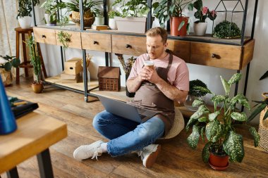 A man with a business shirt sits on floor, engrossed in laptop, surrounded by potted plants in a vibrant plant shop. clipart