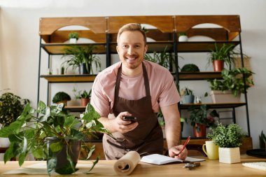 A man in an apron holds a cell phone in a plant shop setting. clipart