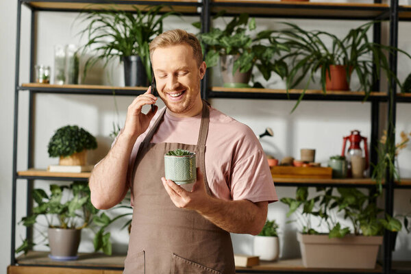 A man in an apron multitasks by holding a plant pot and talking on a cell phone in a botanical shop.