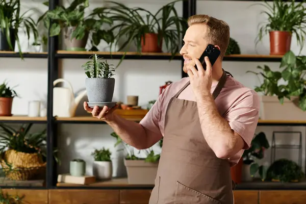 stock image A man multitasking by talking on a cell phone and holding a potted plant in a plant shop setting.