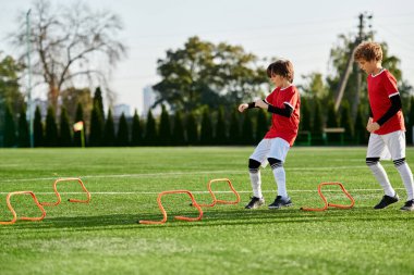 Futbol forması giyen iki genç çocuk çimlerin üzerinde enerjik bir şekilde futbol oynuyor. Pas veriyorlar, top sürüyorlar ve gol atmaya çalışıyorlar. Arka planda, diğer çocuklar onlara tezahürat yapıyor..